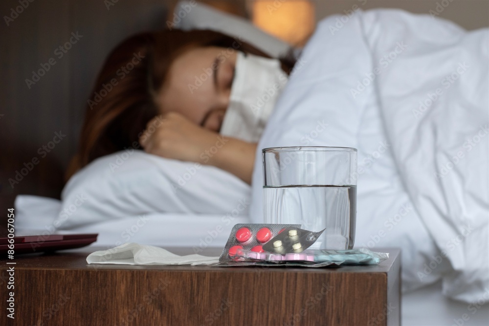 An Asian young woman wearing a mask, appears ill. Selective focus on the pills and glass. An Asian young woman wearing a mask, appears ill. Selective focus on the pills and glass.