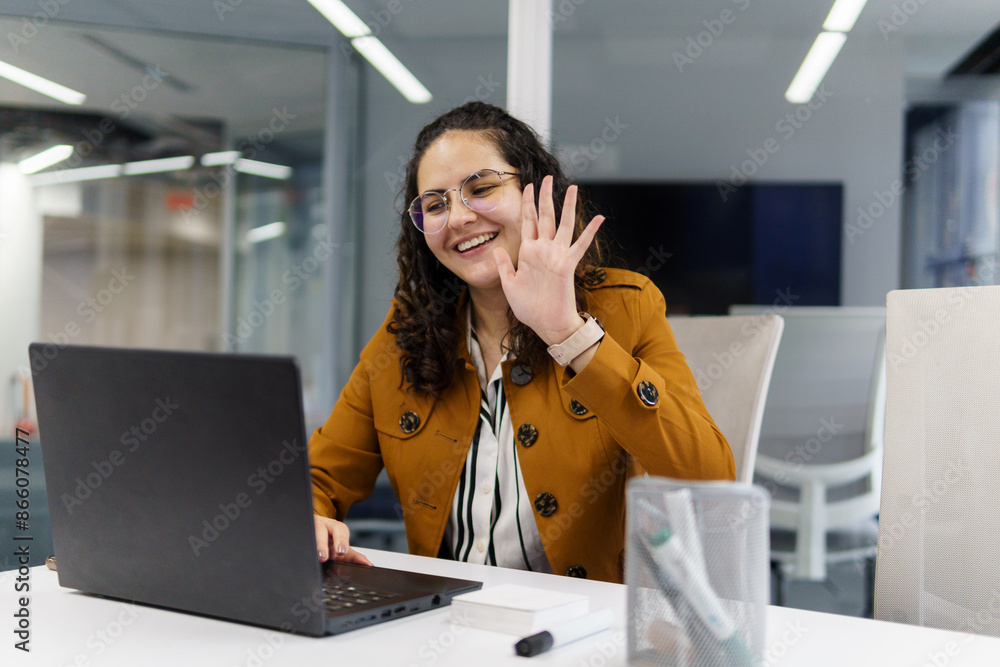 Obraz premium A young, beautiful woman worker in an office who is waving in front of her computer screen, with her hand outstretched and smiling in a yellow coat