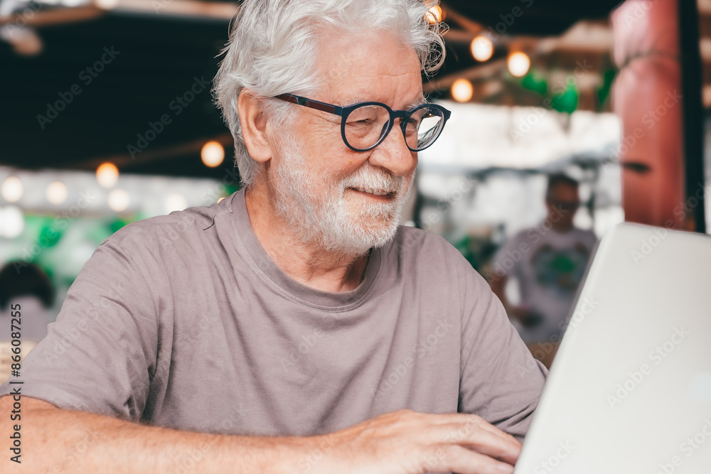 Attractive senior man sitting in a cafe smiling while using laptop. Cheerful senior male, using modern technology expressing happiness and contentment in a relaxed atmosphere Attractive senior man sitting in a cafe smiling while using laptop. Cheerful senior male, using modern technology expressing happiness and contentment in a relaxed atmosphere