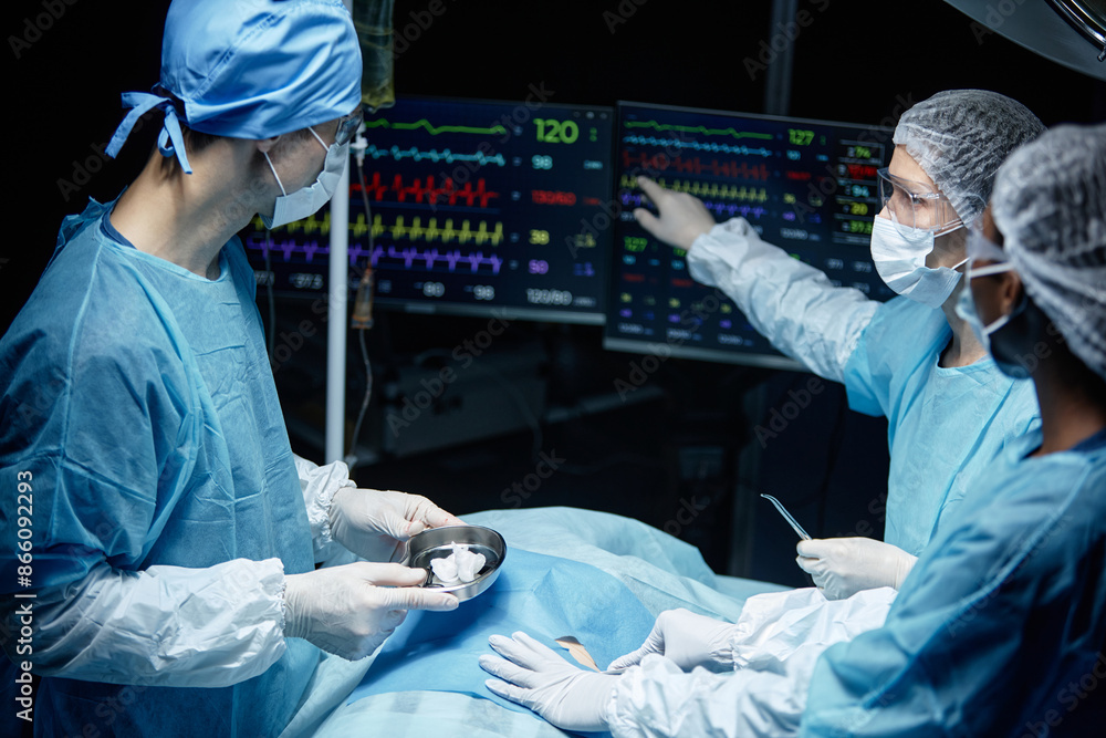 © Seventyfour - Side view portrait of surgical team performing operation in blue lights and looking at monitors with heart rate