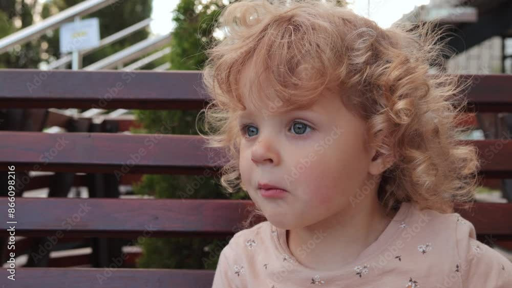 Cute blond wavy haired baby girl sitting on city bench and eating snack while resting outdoor looking around with curious face Cute blond wavy haired baby girl sitting on city bench and eating snack while resting outdoor looking around with curious face