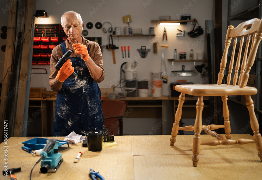 An elderly man preparing to restore an old wooden chair, inserting a drill into drill, restoration and DIY concept. The man is wearing an apron and work gloves, there are tools on the table An elderly man preparing to restore an old wooden chair, inserting a drill into drill, restoration and DIY concept. The man is wearing an apron and work gloves, there are tools on the table