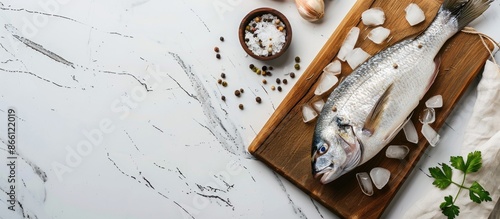 A gutted sea bream or dorado fish lies uncooked on a wooden cutting board with ice, salt, and pepper on a white marble background, ideal for food preparation with space for text. image with copy space photo