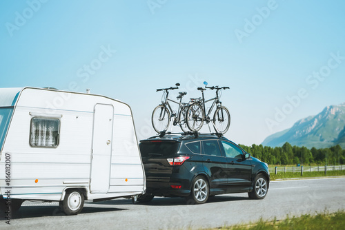 Car with bicycles mounted on top towing a caravan on a sunny highway against a scenic mountain backdrop, family road trip adventure during vacation photo