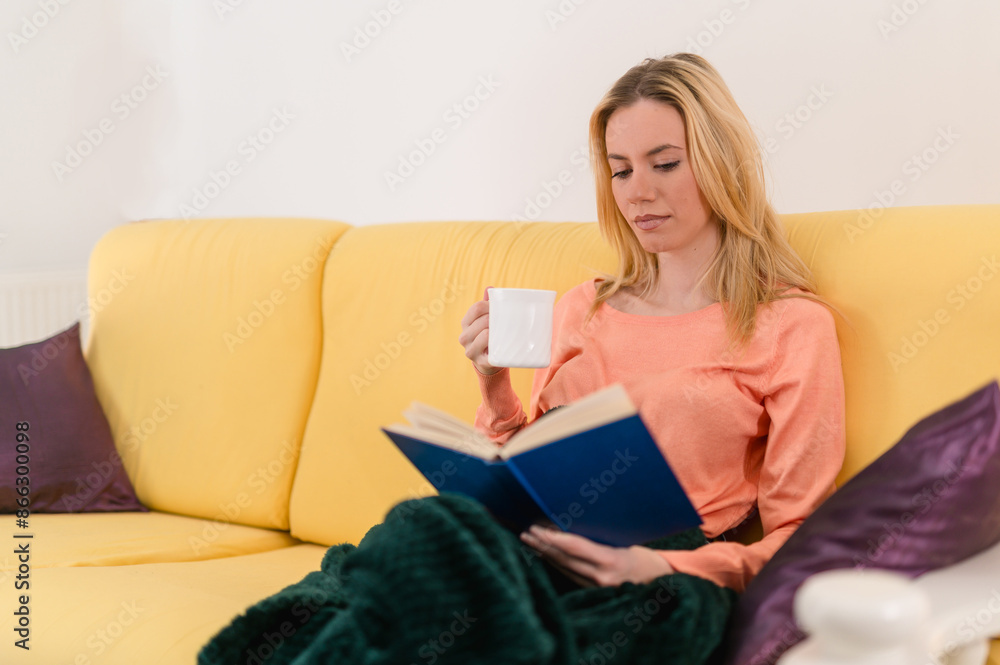 Blonde attractive girl drinking a cup of coffe while immersed in reading of a good novel, sitting on her cozy sofa Blonde attractive girl drinking a cup of coffe while immersed in reading of a good novel, sitting on her cozy sofa
