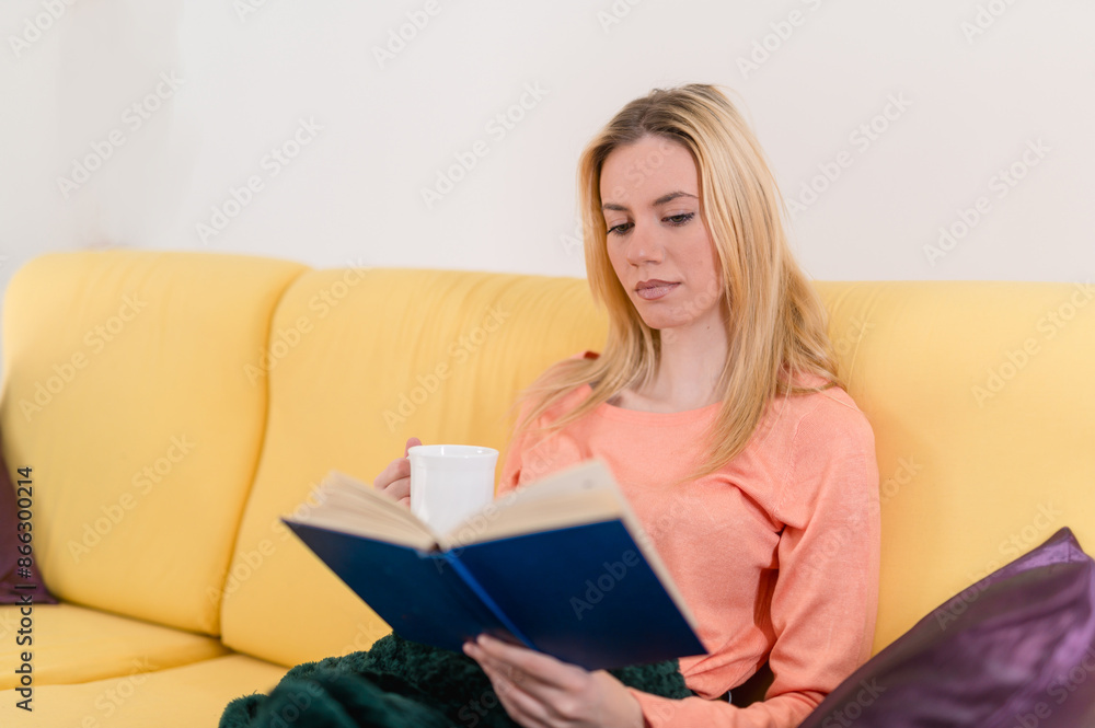 Close shot of a blonde cute girl relaxing, reading a good book and drinking a cup of tea in coziness of her appartment Close shot of a blonde cute girl relaxing, reading a good book and drinking a cup of tea in coziness of her appartment