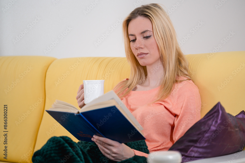 Close shot of a blonde cute girl relaxing, reading a good book and drinking a cup of tea in coziness of her appartment Close shot of a blonde cute girl relaxing, reading a good book and drinking a cup of tea in coziness of her appartment
