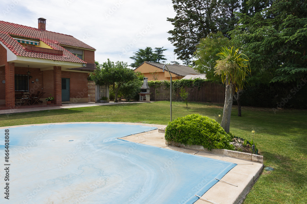 Fototapeta premium Pool covered with blue tarp in a green garden of a private house on a sunny day. End or beginning of season, fall or spring