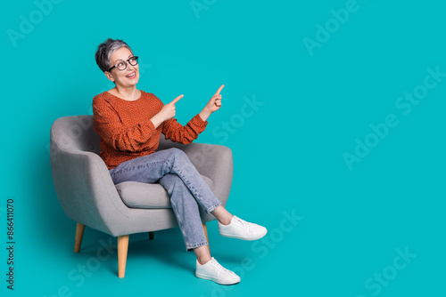 Full length photo of lovely pensioner lady sit armchair look point empty space dressed brown garment isolated on cyan color background