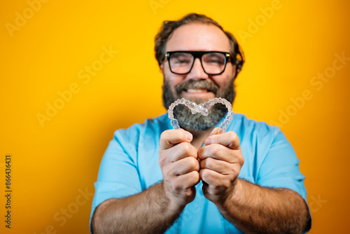 A bearded man wearing a blue shirt and glasses holds a clear retainer in a heart shape. He is smiling and looking directly at the camera. The background is bright yellow. photo