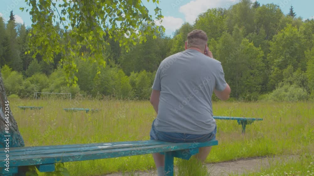 A middle-aged man talks on a mobile phone while sitting on a bench on a sunny summer day. View from the back. A middle-aged man talks on a mobile phone while sitting on a bench on a sunny summer day. View from the back.
