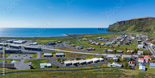 A stunning aerial view of Vik i Myrdal, a coastal town in Iceland. The town features a black sand beach, a small river, green hills, and a large cliff in the background. photo