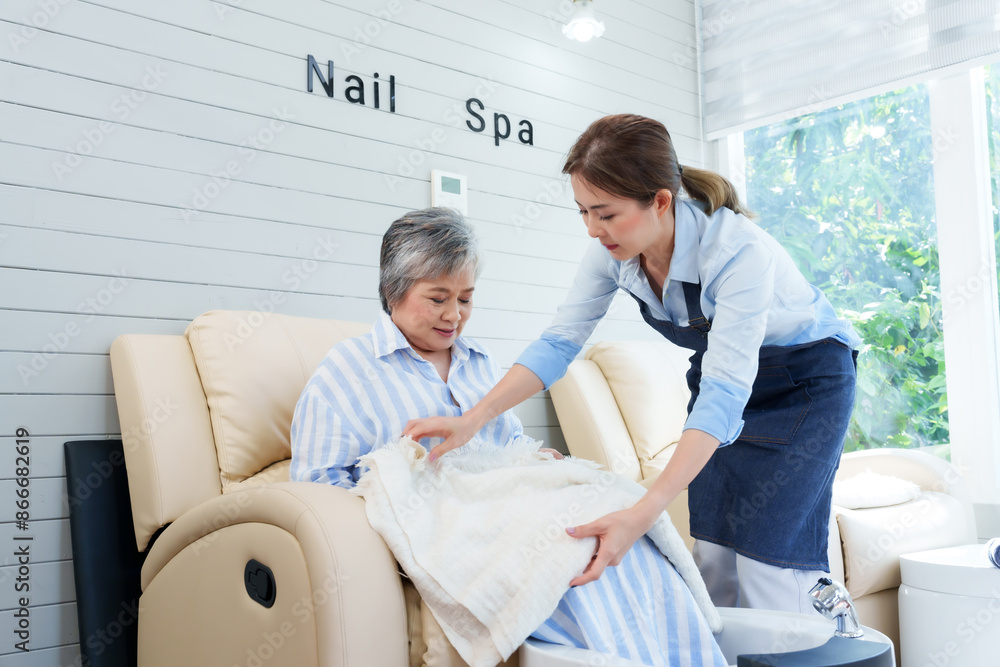 Obraz premium Asian female foot spa shop owner placing blanket on elderly woman in nail spa. Attentive, caring service in bright, cozy environment. client comfort and satisfaction in small business setting.