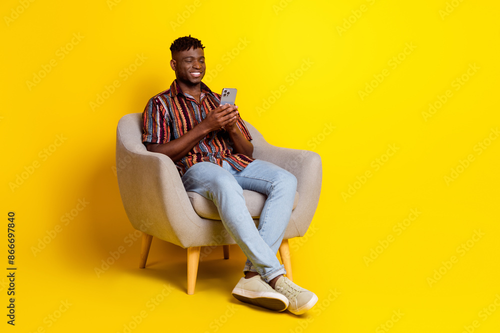 Full body portrait of nice young man sit chair use phone empty space wear shirt isolated on yellow color background Full body portrait of nice young man sit chair use phone empty space wear shirt isolated on yellow color background