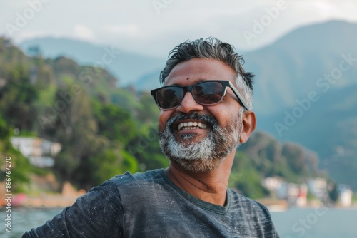 Portrait of a smiling indian man in his 40s wearing a trendy sunglasses while standing against serene lakeside view
