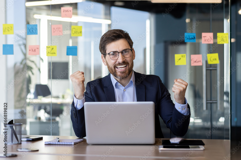 Happy businessman wearing suit celebrating success at office desk with laptop. Concept of achievement, motivation, business success. Office setting with sticky notes on glass wall in background. Happy businessman wearing suit celebrating success at office desk with laptop. Concept of achievement, motivation, business success. Office setting with sticky notes on glass wall in background.