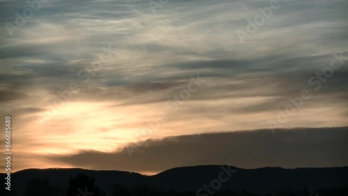 Cloudy sky with moving clouds, pink, orange and yellow colors, and sunlight during sunrise over dark horizon with trees and Silesian Beskid mountains in Poland in December - timelapse.