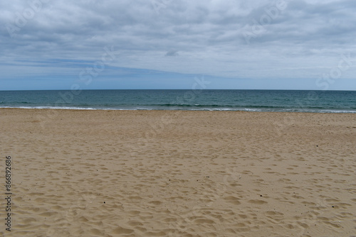 Wide shot of empty beach, sand on the shore, calm sea in the distance, cloudy sky