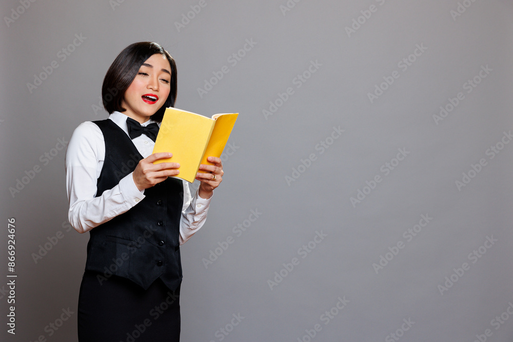 Asian waitress in uniform reading novel during break at restaurant. Catering service young attractive woman employee holding book and enjoying literature with surprised face expression Asian waitress in uniform reading novel during break at restaurant. Catering service young attractive woman employee holding book and enjoying literature with surprised face expression
