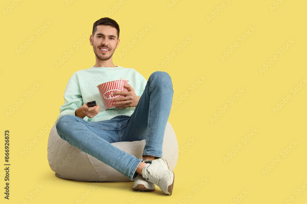 Handsome young happy man with bucket of popcorn watching TV on beanbag chair against yellow background Handsome young happy man with bucket of popcorn watching TV on beanbag chair against yellow background