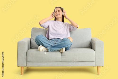 Beautiful young woman in headphones resting on sofa and listening music against yellow background © Pixel-Shot