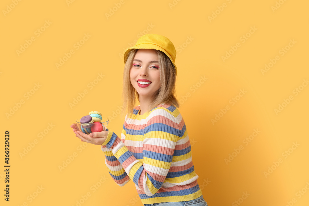 Happy young woman holding glass bowl with sweet macaroons on yellow background Happy young woman holding glass bowl with sweet macaroons on yellow background