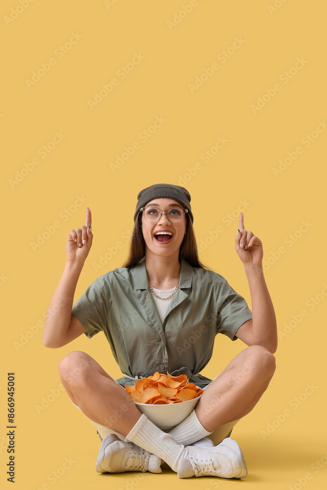 Young woman with bowl of potato chips pointing at something on yellow background Young woman with bowl of potato chips pointing at something on yellow background