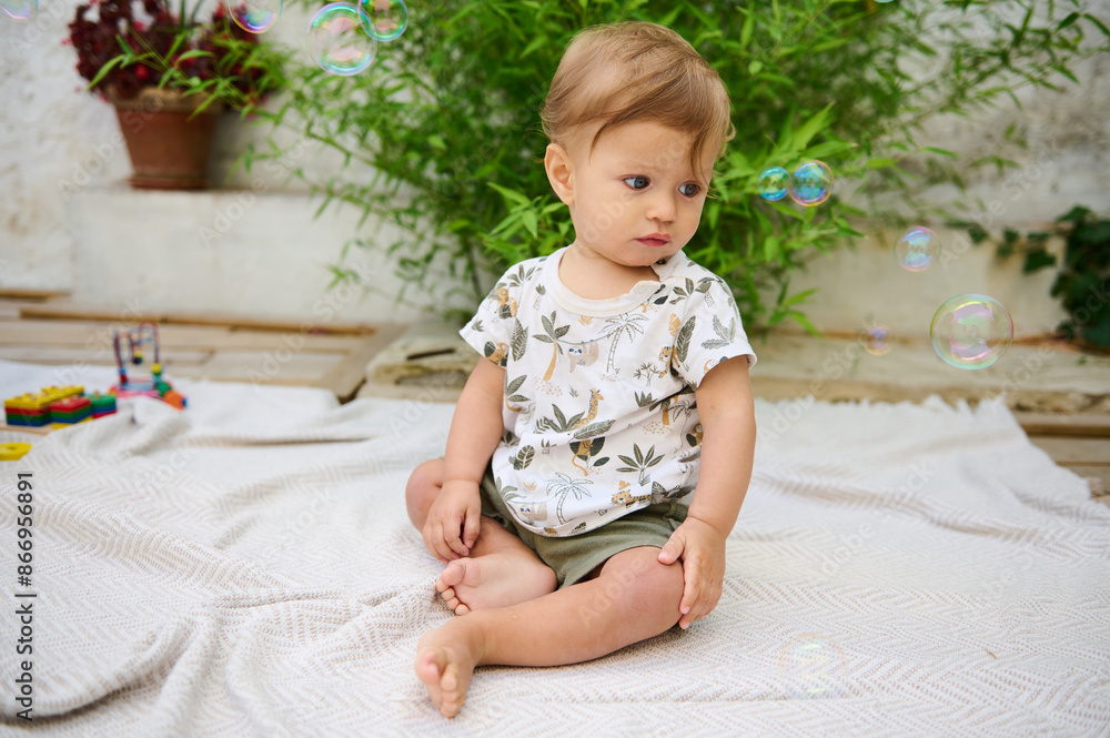 Cute child, a baby boy 10 months old showing interest to flying soap bubbles, sitting on a beige linen blanket outdoors in the patio, surrounded by green plants. Kids. Infancy and babyhood concept Cute child, a baby boy 10 months old showing interest to flying soap bubbles, sitting on a beige linen blanket outdoors in the patio, surrounded by green plants. Kids. Infancy and babyhood concept