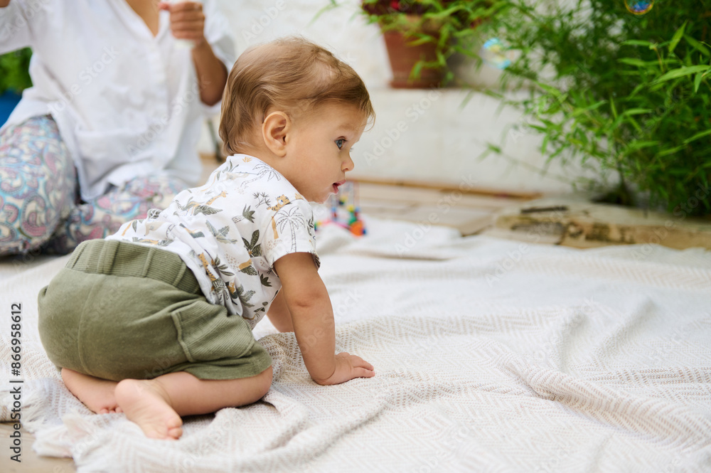 Adorable Caucasian redhead baby boy crawling on a beige linen blanket outdoors. Green bamboo leaves and loving mum blowing soap bubbles, can being seen on the blurred background. Infancy and babyhood Adorable Caucasian redhead baby boy crawling on a beige linen blanket outdoors. Green bamboo leaves and loving mum blowing soap bubbles, can being seen on the blurred background. Infancy and babyhood