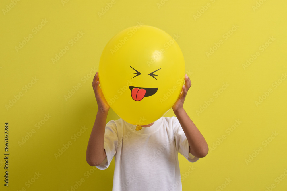 Tongue emoji. Cute Asian boy hiding his face behind balloon emoticon sticking out tongue isolated on yellow background. Teasing emoji with closed eyes Tongue emoji. Cute Asian boy hiding his face behind balloon emoticon sticking out tongue isolated on yellow background. Teasing emoji with closed eyes