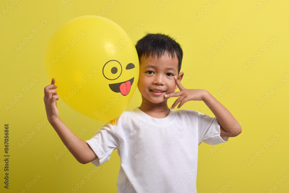 Tongue emoji. Cute Asian boy holding tongue out emoticon balloon isolated on yellow background. Teasing emoji with closed eyes