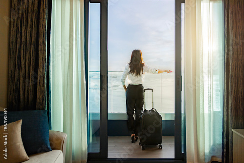 Confident business leader with suitcase solo travel, standing and enjoying large balcony, looking away on tropical paradise beach during sunset time and enjoys her vacation. Colorful sunset over ocean