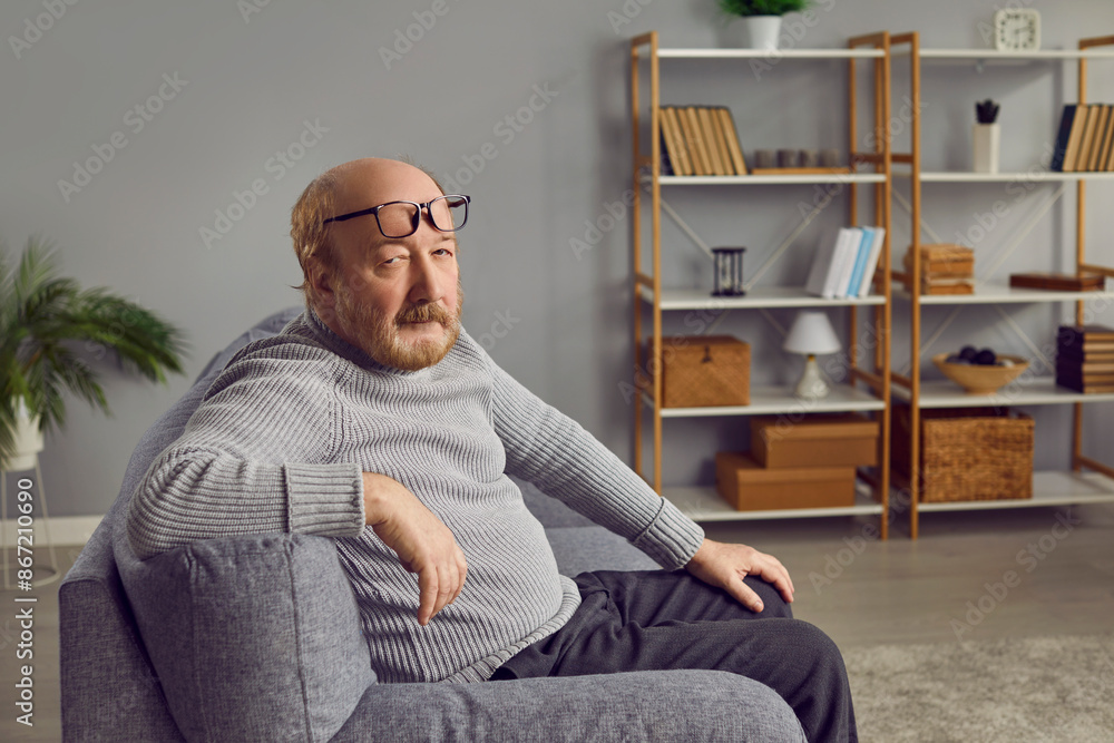 Fototapeta premium Portrait of a senior man at home. Retired old bald man in a gray sweater, with glasses on his forehead, sitting on the sofa in the living room and looking at the camera with a serious face expression