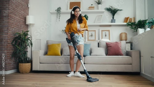 Video of young happy woman listening and dancing to music while cleaning the living room floor with a vaccum cleaner