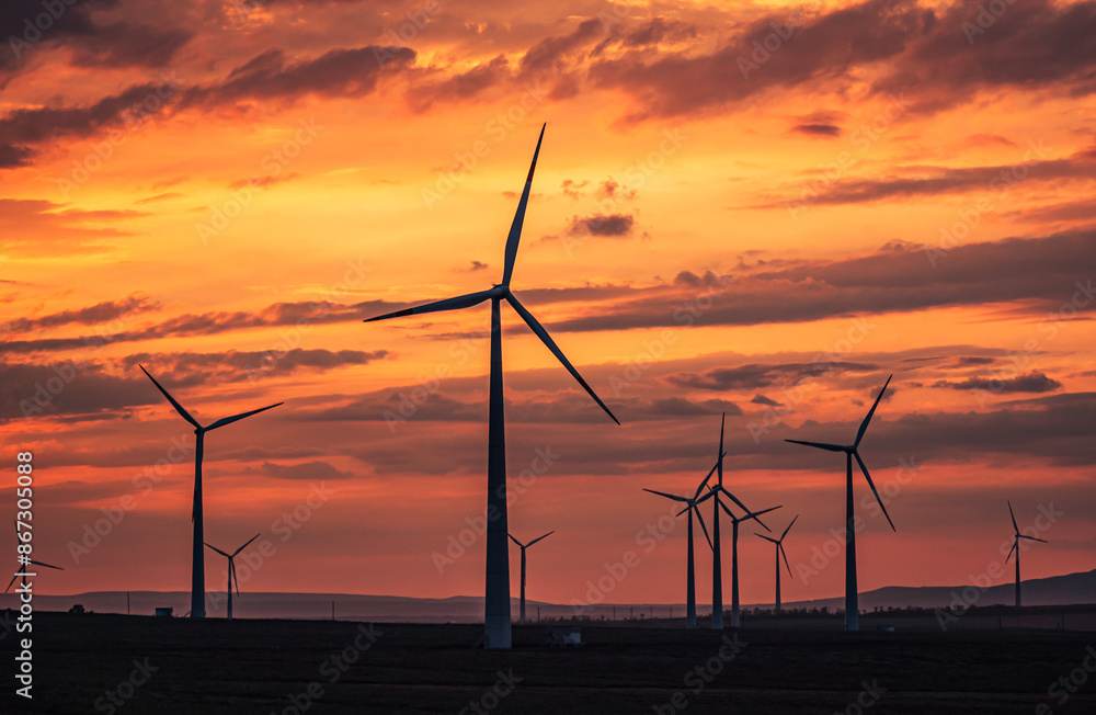 Fototapeta premium Multiple wind turbines silhouetted against a vibrant sunset sky. The turbines stand tall against the backdrop of orange and red clouds.