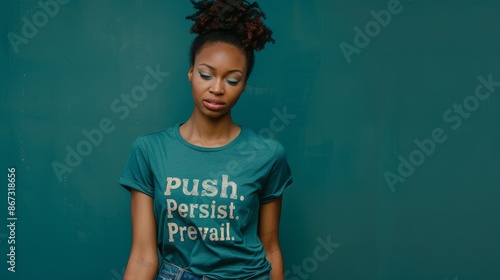 A young woman wearing a teal t-shirt with the words Push. Persist. Prevail. printed on it stands confidently in front of a teal backdrop