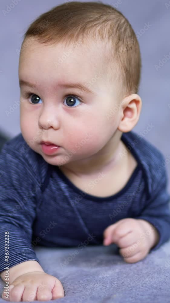 Adorable Caucasian boy in blue clothes lies on belly waving his feet. Nice curious toddler looks up with interest sweetly rising his eyebrows. Vertical video Adorable Caucasian boy in blue clothes lies on belly waving his feet. Nice curious toddler looks up with interest sweetly rising his eyebrows. Vertical video