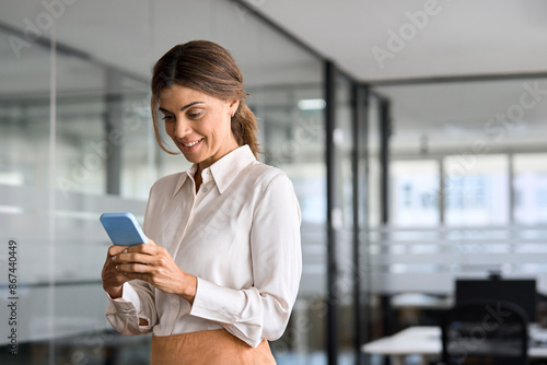 Happy busy middle aged business woman executive looking at cellphone with mobile phone in hands. Smiling mature female bank manager entrepreneur using smartphone standing in office at work. Copy space