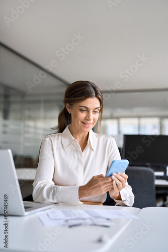 Vertical photo of busy happy professional middle aged business woman using mobile phone working in office. Smiling mature executive holding cellphone, female employee making payments on smartphone.