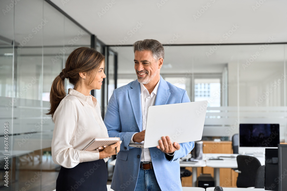 Happy professional smiling businessman and businesswoman having conversation at work. Executives team of two business people working talking using laptop technology standing in corporate office. Happy professional smiling businessman and businesswoman having conversation at work. Executives team of two business people working talking using laptop technology standing in corporate office.