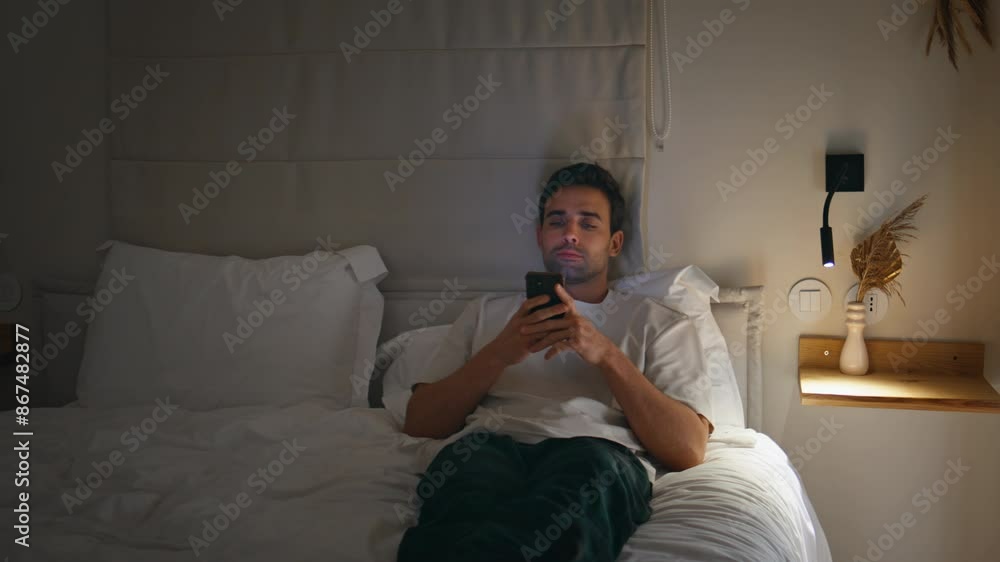 Sleepless man browsing phone in bedroom lit by lamp light. Guy using smartphone Sleepless man browsing phone in bedroom lit by lamp light. Guy using smartphone