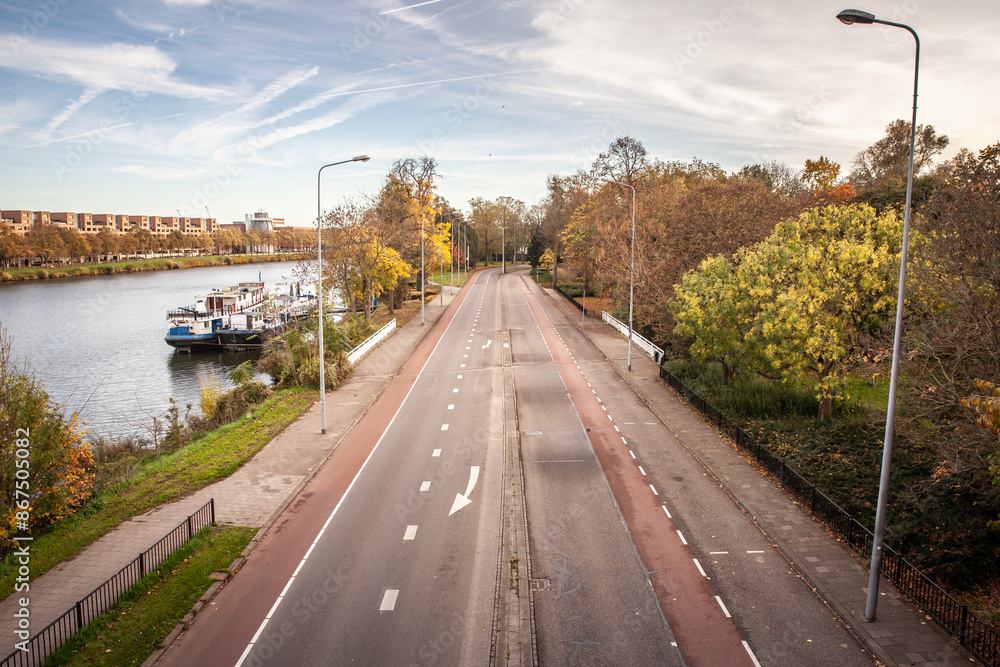 Obraz premium A four-lane street runs alongside the Maas River in Maastricht, with dedicated bicycle lanes and minimal traffic. The photo captures the blend of urban infrastructure and natural elements. The tree-li