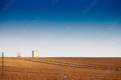 Picture of a plowed field with furrows on a sunny afternoon with blue sky in serbia, in deliblatska pescara with a silo in background. This image features an expansive agricultural field with silos in