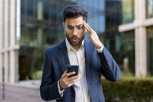 Fotografie Shocked businessman in blue suit looking at smartphone with surprised expression outside modern office building, feeling shocked, stressed, and concerned while reading unexpected news or error