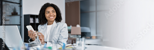 Young African American business manager using cell phone mobile app, laptop. Latin smiling adult businesswoman in suit holding smartphone sitting at office table, working online. Banner, copy space photo