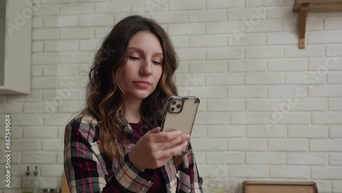 A woman is seen using a smartphone in a cozy kitchen while wearing a plaid shirt