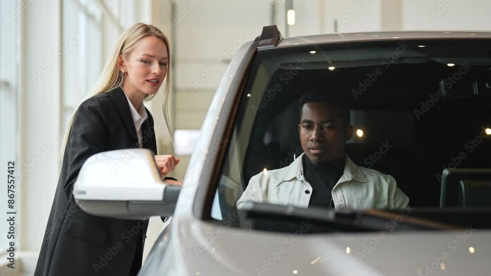 Friendly female auto dealer in business suit having communication with African-American male customer sitting in car salon. Professional blonde saleswoman helping man client to choose best automobile. Friendly female auto dealer in business suit having communication with African-American male customer sitting in car salon. Professional blonde saleswoman helping man client to choose best automobile.