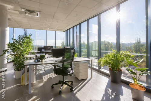 A stylish, minimalist office space with large windows, sleek furniture, and vibrant indoor plants. The scene is brightly lit by natural sunlight, creating an inviting workspace photo