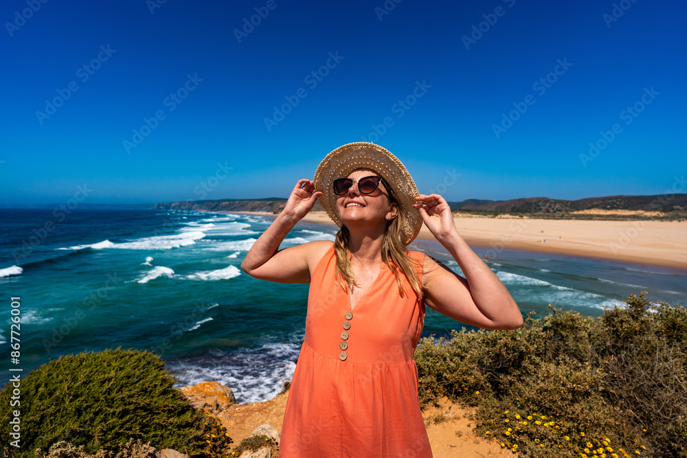 Beautiful woman in fashionable orange dress at viewpoint on cliff near sunny and sandy beach Bordeira, Algarve, Portugal Beautiful woman in fashionable orange dress at viewpoint on cliff near sunny and sandy beach Bordeira, Algarve, Portugal