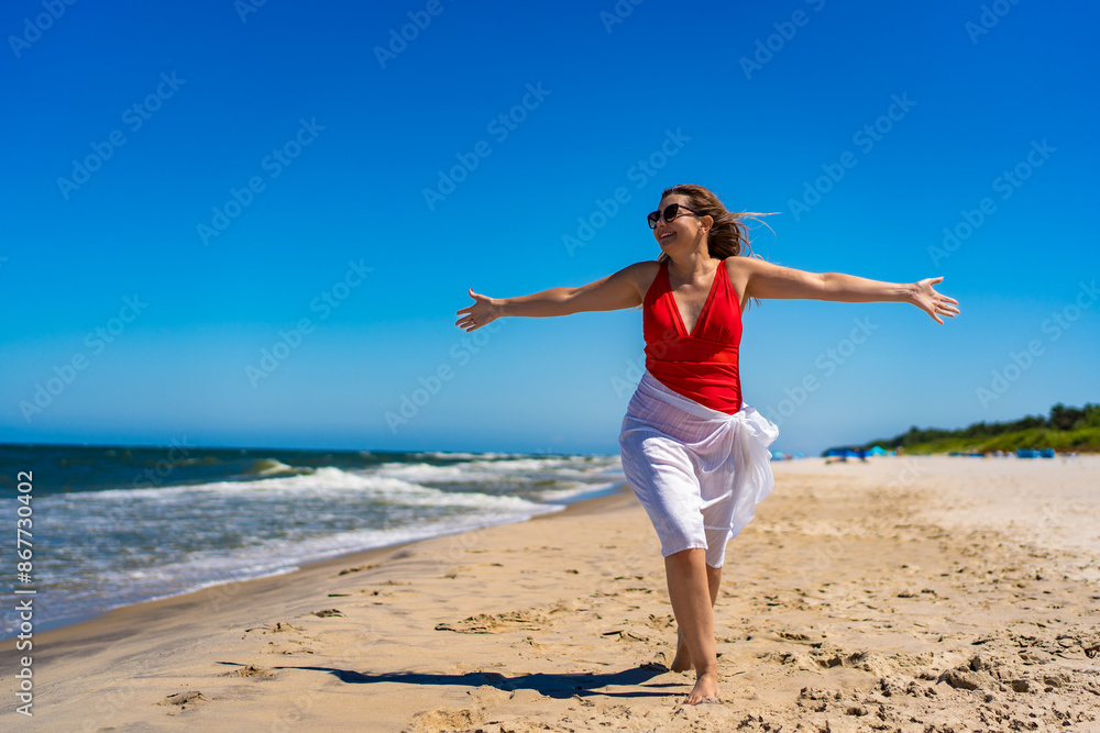 Summer vacation. Beautiful young woman in red swimsuit and white scarf and sunglasses walking with her arms spread out on the sandy beach on a beautiful sunny day Summer vacation. Beautiful young woman in red swimsuit and white scarf and sunglasses walking with her arms spread out on the sandy beach on a beautiful sunny day